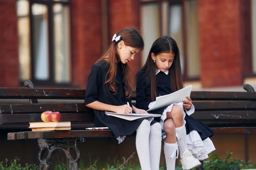 Two schoolgirls is sitting outside together near school building.