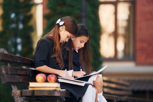 Sitting and reading. Two schoolgirls is outside together near school building.
