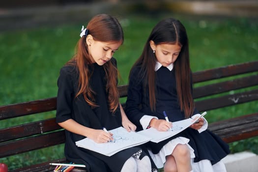 Sitting and reading. Two schoolgirls is outside together near school building.