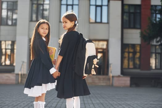 Walking together. Two schoolgirls is outside near school building.