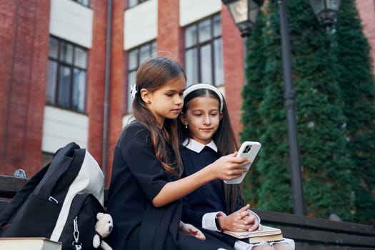 Sitting together. Two schoolgirls is outside near school building.