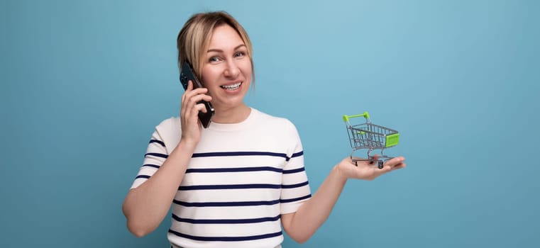panoramic photo of young cute casual woman shopaholic talking on phone holding empty shopping cart in supermarket on blue background with copy space.