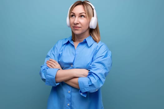 charming european girl in a casual shirt listens to music in big white headphones on a blue isolated background.