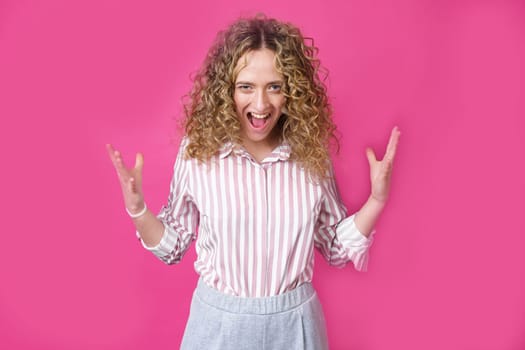 A curly-haired woman raises her palms in joy, glad to receive an amazing gift from someone, screams loudly, wearing a striped shirt. Isolated on a purple background.