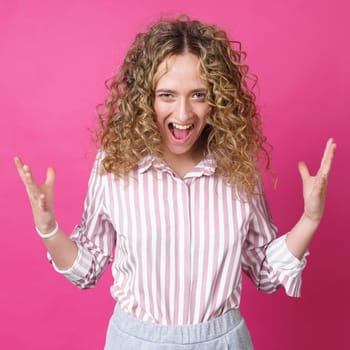 A curly-haired woman raises her palms in joy, glad to receive an amazing gift from someone, screams loudly, wearing a striped shirt. Isolated on a purple background.