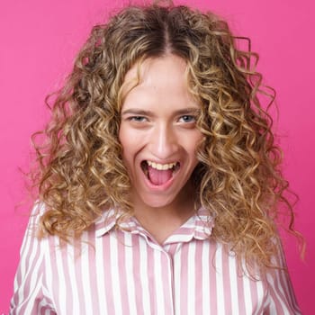 A curly-haired woman raises her palms in joy, glad to receive an amazing gift from someone, screams loudly, wearing a striped shirt. Isolated on a purple background.