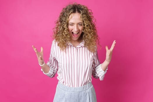 A curly-haired woman raises her palms in joy, glad to receive an amazing gift from someone, screams loudly, wearing a striped shirt. Isolated on a purple background.