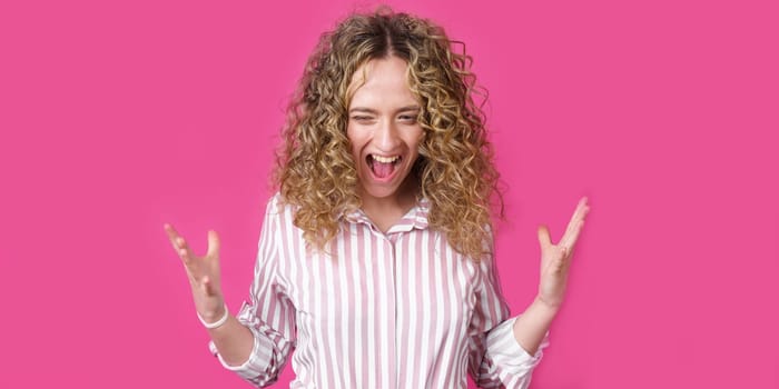 A curly-haired woman raises her palms in joy, glad to receive an amazing gift from someone, screams loudly, wearing a striped shirt. Isolated on a purple background.