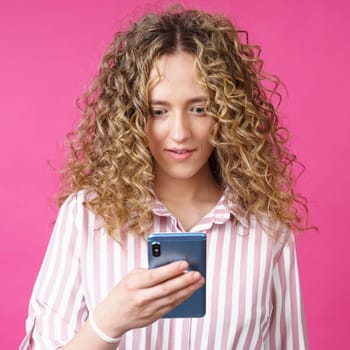 A fashionable woman in a striped shirt, holds a mobile phone, gasps in surprise, reads amazing news. Isolated on pink background