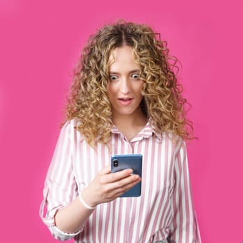 A fashionable woman in a striped shirt, holds a mobile phone, gasps in surprise, reads amazing news. Isolated on pink background