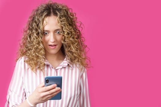 A fashionable woman in a striped shirt, holds a mobile phone, gasps in surprise, reads amazing news. Isolated on pink background