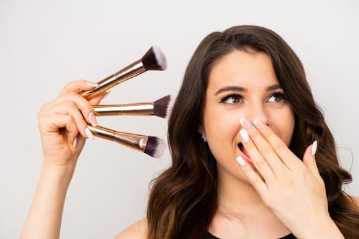 Beautiful brunette woman holding makeup brushes covering her mouth with a hand with a surprised expression on the grey background