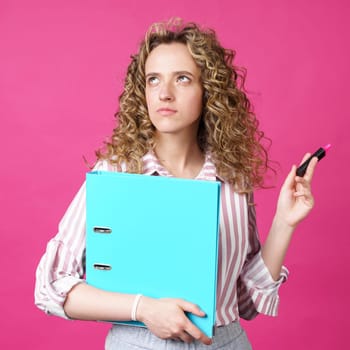 Fashionable woman in a striped shirt holding a folder with documents and a marker. Looks up. Isolated on pink background