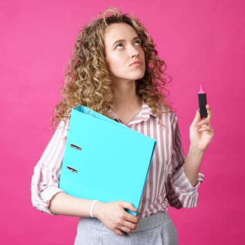 Fashionable woman in a striped shirt holding a folder with documents and a marker. Looks up. Isolated on pink background