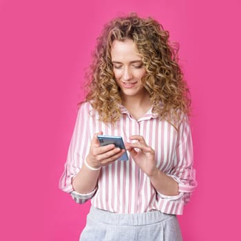 Fashionable woman in a striped shirt, typing a message on the phone and smiling. Isolated on pink background