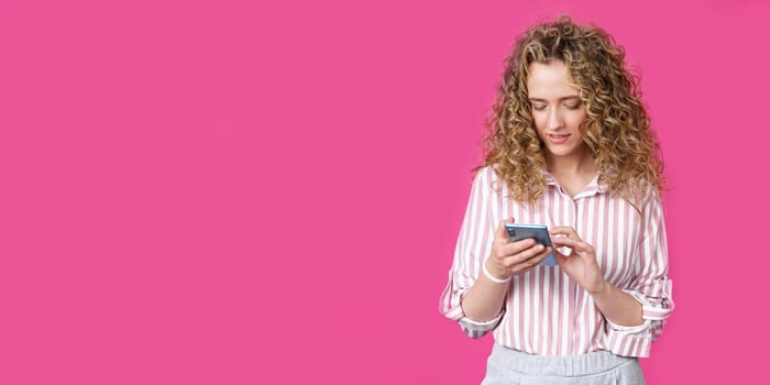 Fashionable woman in a striped shirt, typing a message on the phone and smiling. Isolated on pink background