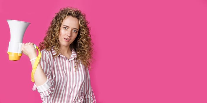 A young woman holds a megaphone in her hands and looks at the camera. Isolated pink background. People sincere emotions lifestyle concept.