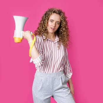 A young woman holds a megaphone in her hands and looks at the camera. Isolated pink background. People sincere emotions lifestyle concept.