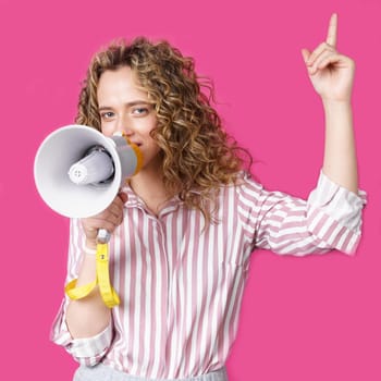 Young woman speaks into a megaphone and raises her finger up. Isolated pink background. People sincere emotions lifestyle concept.