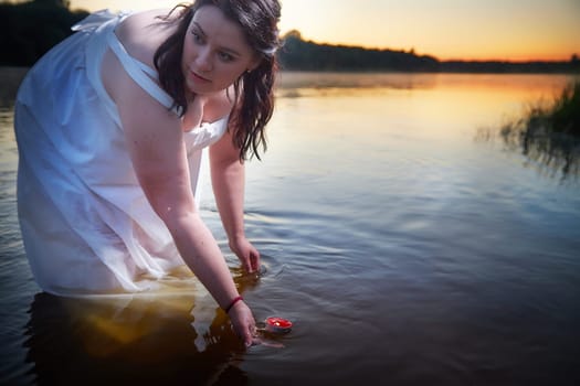 Slavic plump plump chubby girl in long white dress on the feast of Ivan Kupala with flowers and water in a river or lake on summer evening