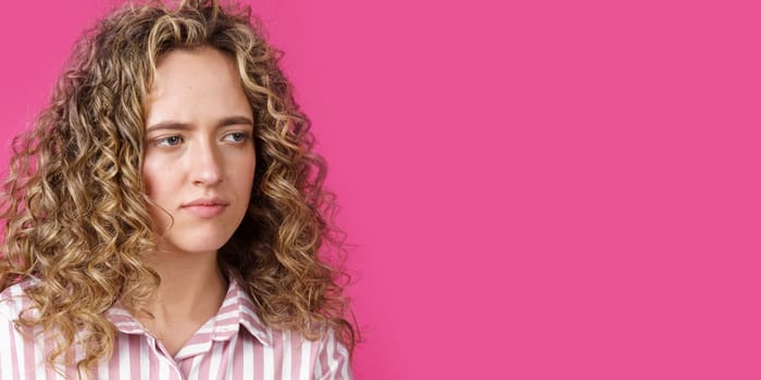 A side shot of a beautiful upset woman with curly hair holding her arms folded. Isolated on pink background.