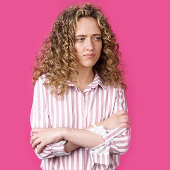 A side shot of a beautiful upset woman with curly hair holding her arms folded. Isolated on pink background.