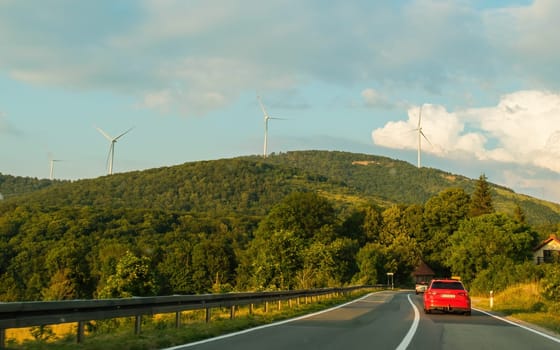 Red car drives on road against green hillside with windmill farm. Producing ecological electrical energy among green landscape under blue sky