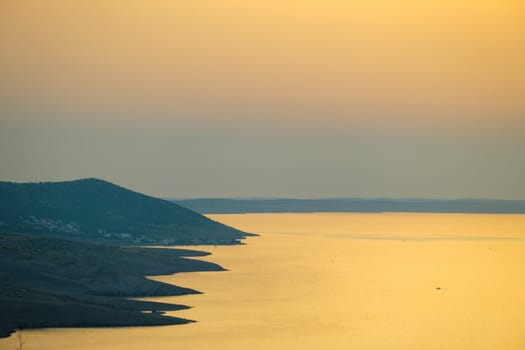 Calm sea surface reflects orange sunlight. Sun sets behind blurry mountains on coast. Dark silhouettes of coastline hills in evening