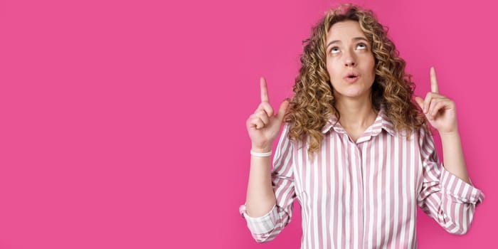 Young beautiful woman, amazed and surprised, looking up and pointing with fingers and raised hands. Isolated on pink background.