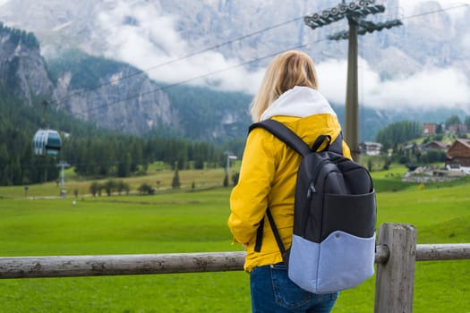 Young woman with blonde hair and yellow jacket looks into the distance of a village in the Dolomites.