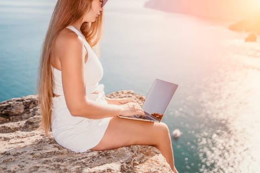 Successful business woman in yellow hat working on laptop by the sea. Pretty lady typing on computer at summer day outdoors. Freelance, travel and holidays concept.