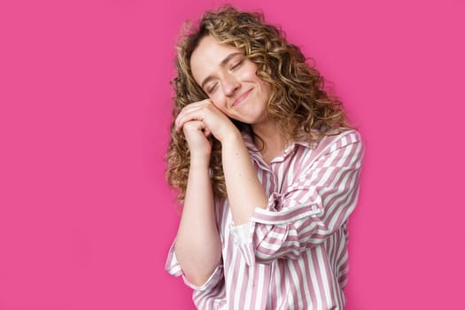 Happy contented woman holds her hands together near the face. Isolated on pink background