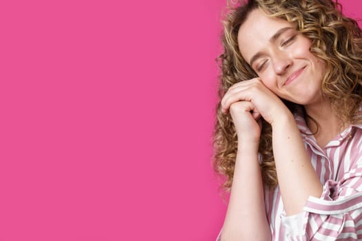 Happy contented woman holds her hands together near the face. Isolated on pink background