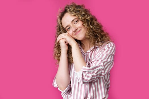 Happy contented woman holds her hands together near the face. Isolated on pink background