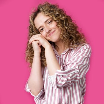 Happy contented woman holds her hands together near the face. Isolated on pink background