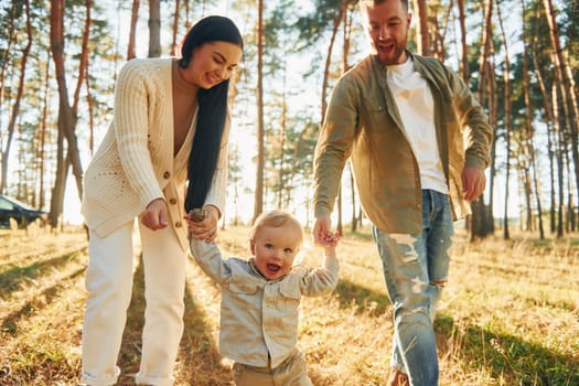 Holding little girl by hands. Happy family of father, mother and little daughter is in the forest.