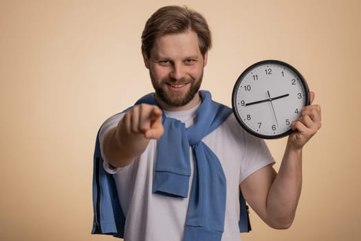 It is your time. Caucasian smiling man showing time on wall office clock, ok, thumb up, approve, pointing finger at camera, hurry up, approve. Young bearded guy isolated on beige studio background