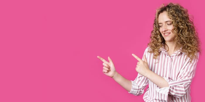 A cheerful woman points to a copy space, discusses an amazing ad, gives way or a direction, wears a striped shirt, has a pleasant smile, and feels optimistic. Isolated on pink background
