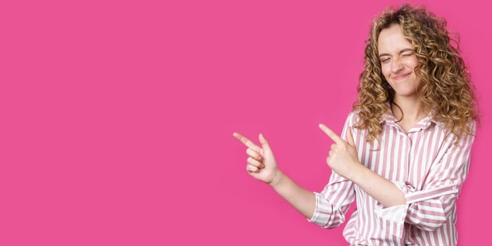 A cheerful woman points to a copy space, discusses an amazing ad, gives way or a direction, wears a striped shirt, has a pleasant smile, and feels optimistic. Isolated on pink background