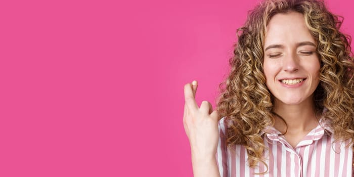 Waiting for a special moment. A portrait of a smiling woman with closed eyes who keeps her fingers crossed. Make a wish. Isolated on a pink background. People sincere emotions concept.