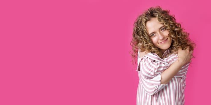Waiting for a special moment. Portrait of a smiling woman who hugs herself with her arms. Isolated on a pink background. People sincere emotions concept.