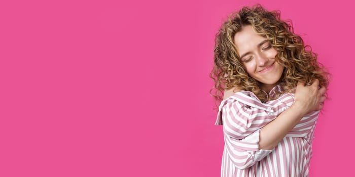 Waiting for a special moment. Portrait of a smiling woman with closed eyes who hugs herself with her arms. Isolated on a pink background. People sincere emotions concept.