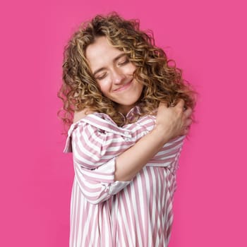 Waiting for a special moment. Portrait of a smiling woman with closed eyes who hugs herself with her arms. Isolated on a pink background. People sincere emotions concept.