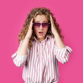 A young woman holds her head with her hands, surprised. Isolated on pink background