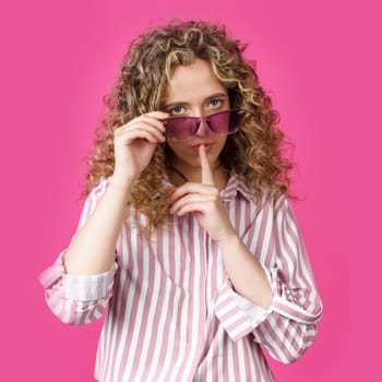 The young woman holds onto her glasses and makes a gesture with her hands - quietly. Secret, secret, isolated on pink background