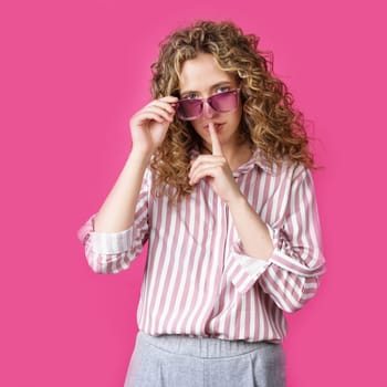 The young woman holds onto her glasses and makes a gesture with her hands - quietly. Secret, secret, isolated on pink background