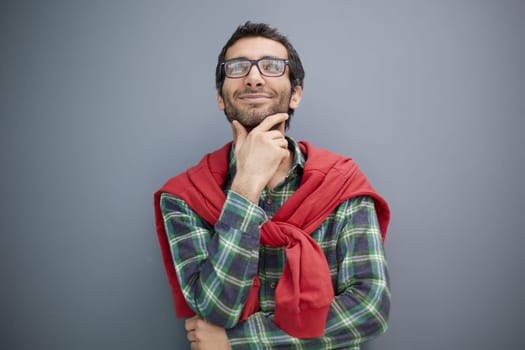 Handsome adult man in elegant casual wear and stylish glasses rubbing his chin and looking away while standing against light gray background