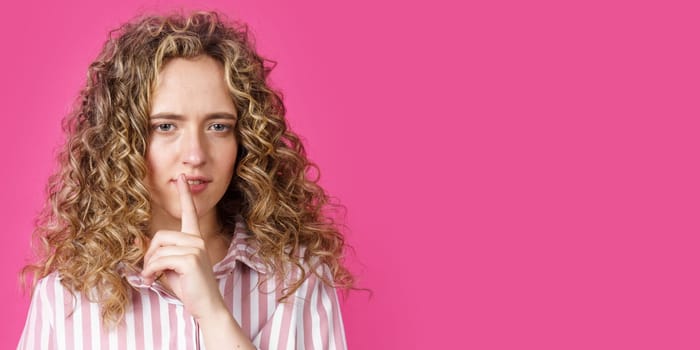 A young woman makes a gesture with her hands quietly, applying them to her lips. Isolated on pink background