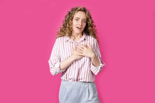 The woman holds her hands on her heart, wearing a striped shirt, expresses gratitude, has a friendly expression on her face. Isolated on pink background