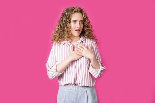 The woman holds her hands on her heart, wearing a striped shirt, expresses gratitude, has a friendly expression on her face. Isolated on pink background